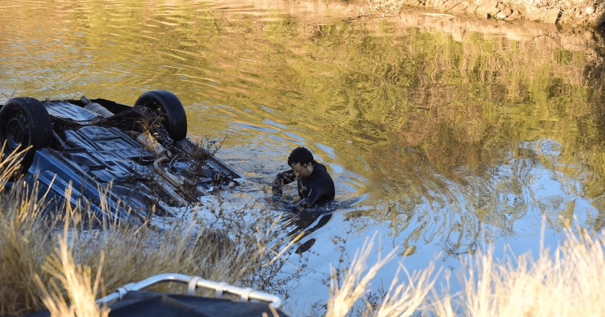 Vehículo volcado en el agua con persona cercana, rodeado de vegetación y reflejos.