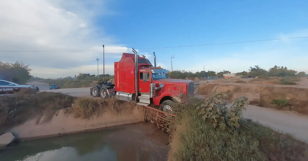 Tráiler rojo embancado en terraplén con remolque sobresaliendo, cielo despejado y vehículos a lo lejos.
