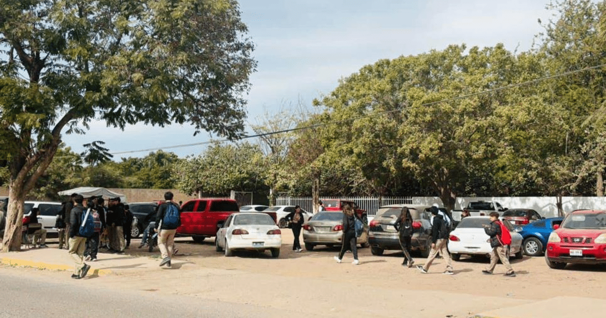 Estudiantes caminando en un estacionamiento escolar en Guasave, Sinaloa, con coches y árboles al fondo.