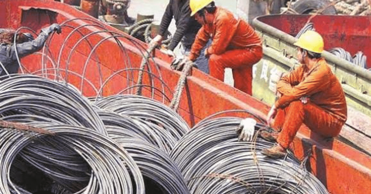 Trabajadores en entorno industrial manipulando rollos de acero, con contenedores y estructuras metálicas de fondo.