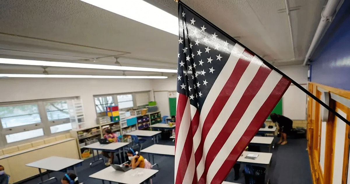 Salón de clases en Estados Unidos con estudiantes y bandera estadounidense en primer plano.