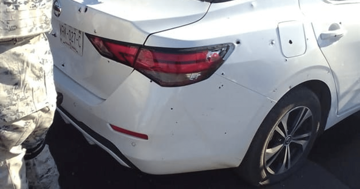 Automóvil blanco dañado por impactos de bala en Culiacán, con un oficial a la izquierda.