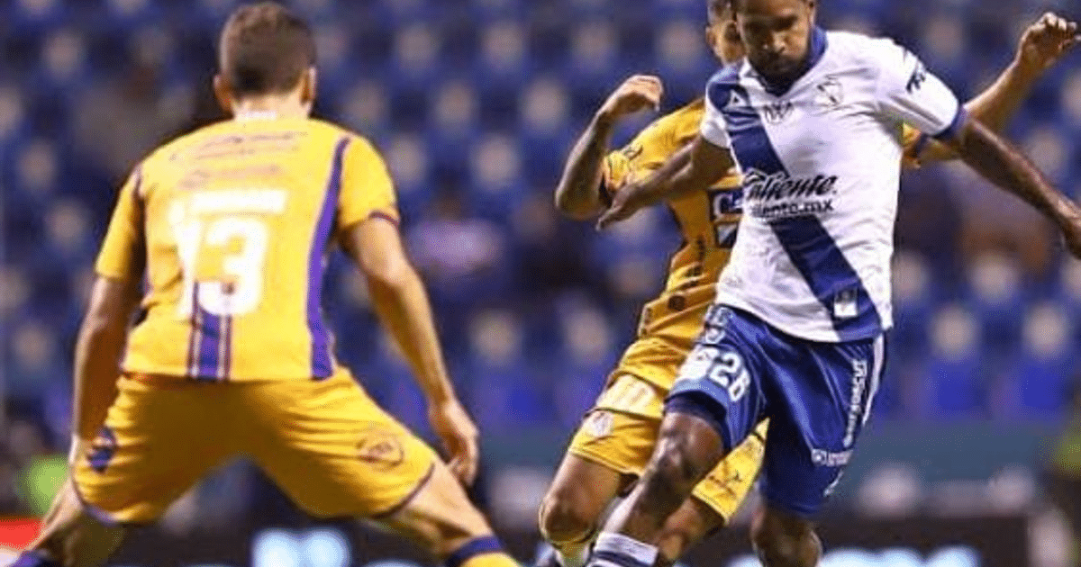 Jugador de fútbol con camiseta blanca y azul pateando el balón en un partido, con otros jugadores y asientos de fondo.