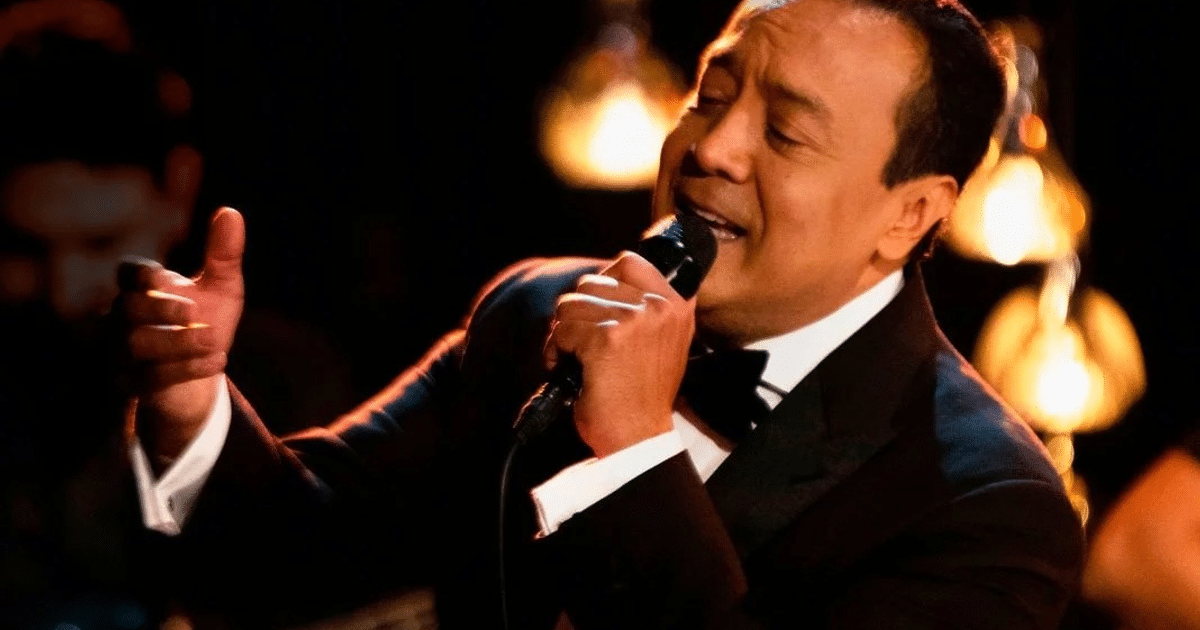 Carlos Cuevas cantando en el escenario con traje formal y luces cálidas de fondo.