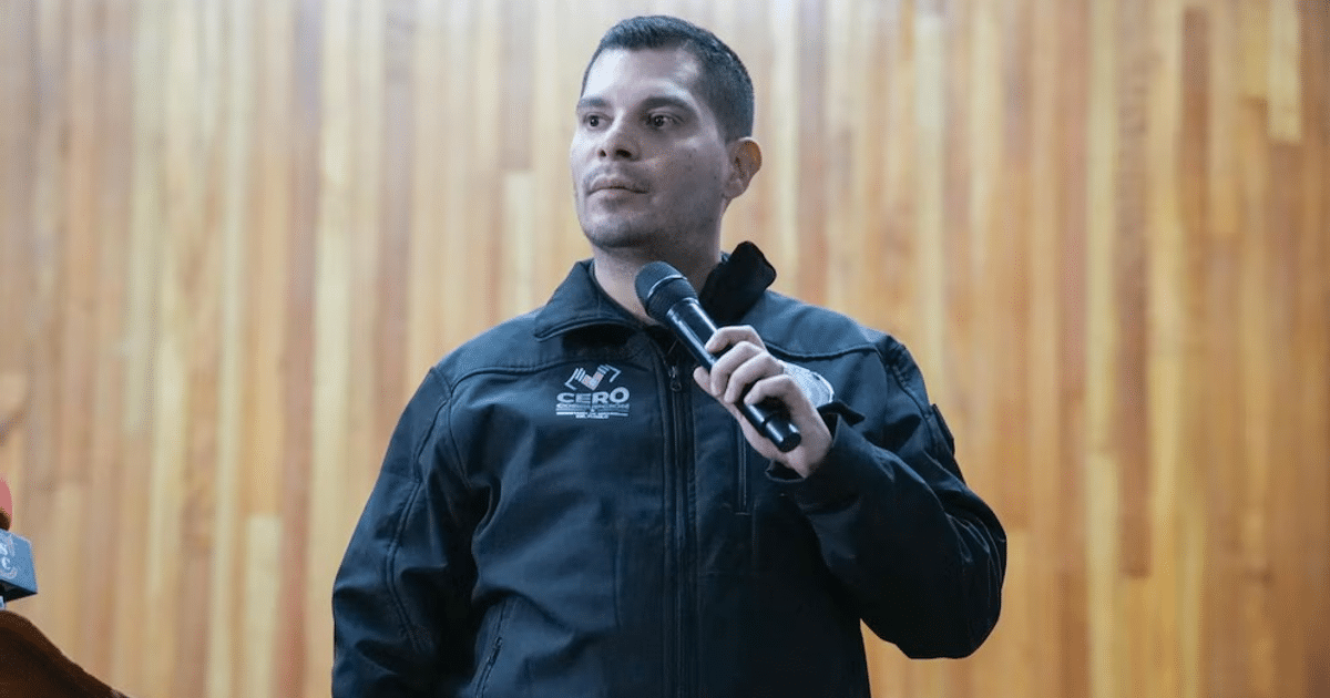 Óscar Aparicio Avendaño hablando en un evento de seguridad en Chiapas, sosteniendo un micrófono y vistiendo un abrigo negro con el logo de 'CERO'.