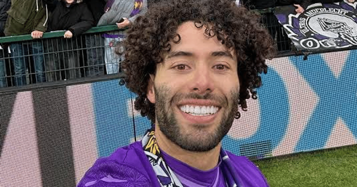 César 'Chino' Huerta sonríe en un selfi tras su debut con el RSC Anderlecht, con una camiseta morada y una bandera al fondo.