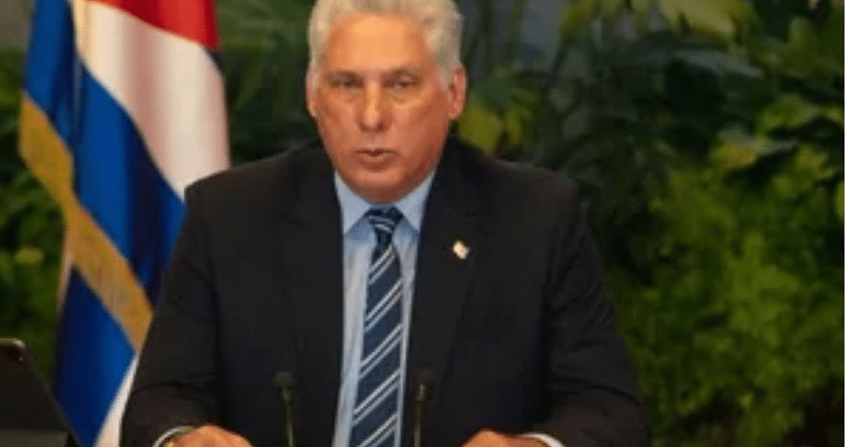 Persona hablando en un evento formal con bandera cubana de fondo