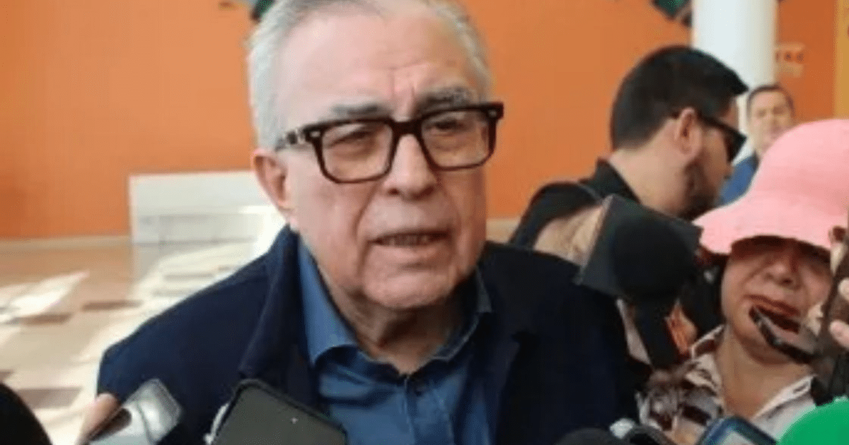 Gobernador de Sinaloa en conferencia de prensa con micrófonos y personas alrededor, fondo naranja.