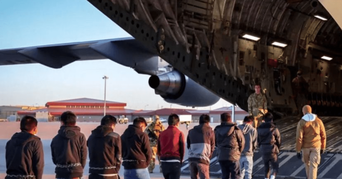Grupo de personas encadenadas escoltadas hacia un avión militar en una pista de aterrizaje