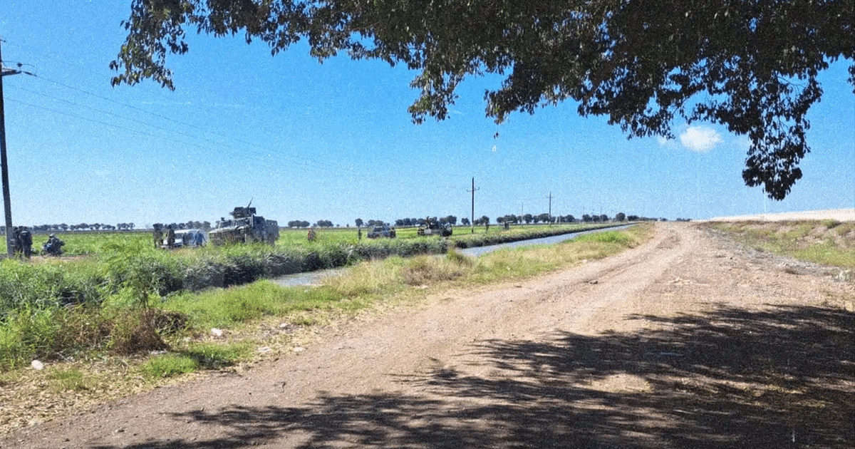 Paisaje rural en Zapotillo con camino de tierra, canal de agua, campos verdes, vehículos y personas a lo lejos, líneas eléctricas y cielo despejado.