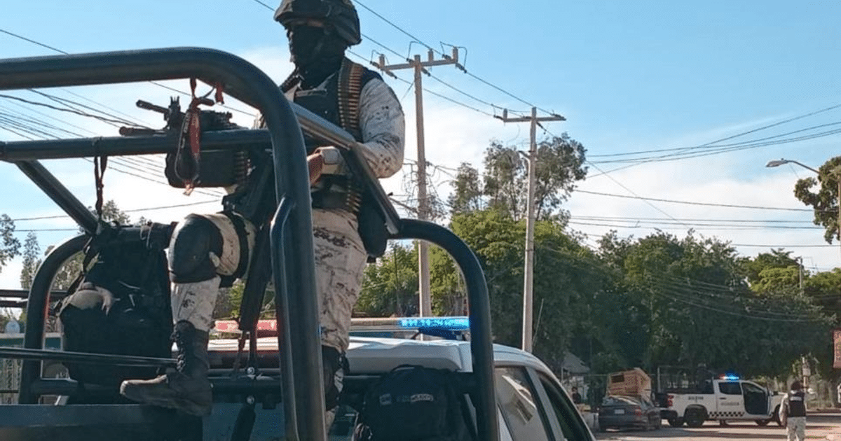 Individuo armado en operativo de seguridad en Culiacán, con vehículos policiales de fondo.