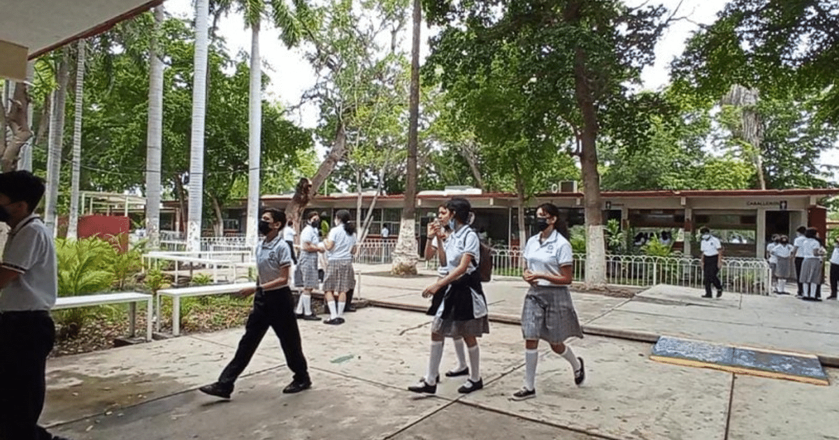 Estudiantes en uniforme y mascarillas socializando al aire libre en un entorno escolar con árboles y edificios al fondo.