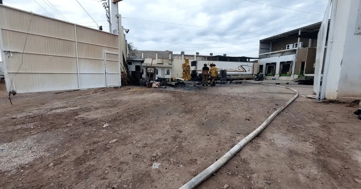 Área afectada por incendio en bodega de aguacates en Culiacán, con portón, personas, vehículos y escombros visibles.