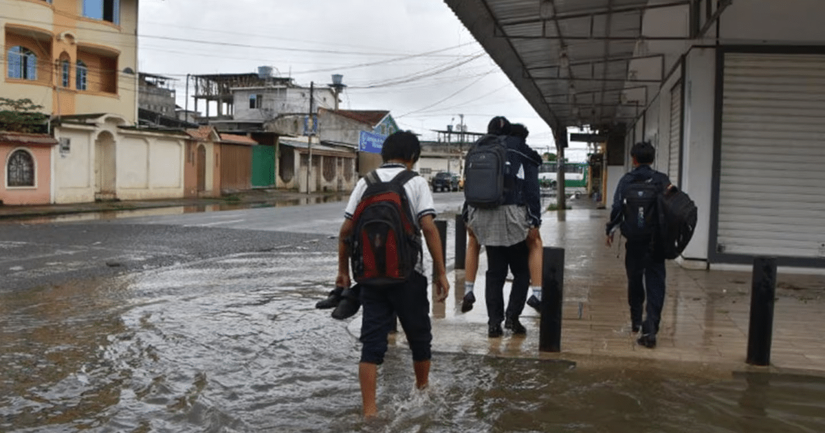 Grupo de jóvenes caminando por una calle inundada en Culiacán, con mochilas y uno cargando a otro, en un día nublado.