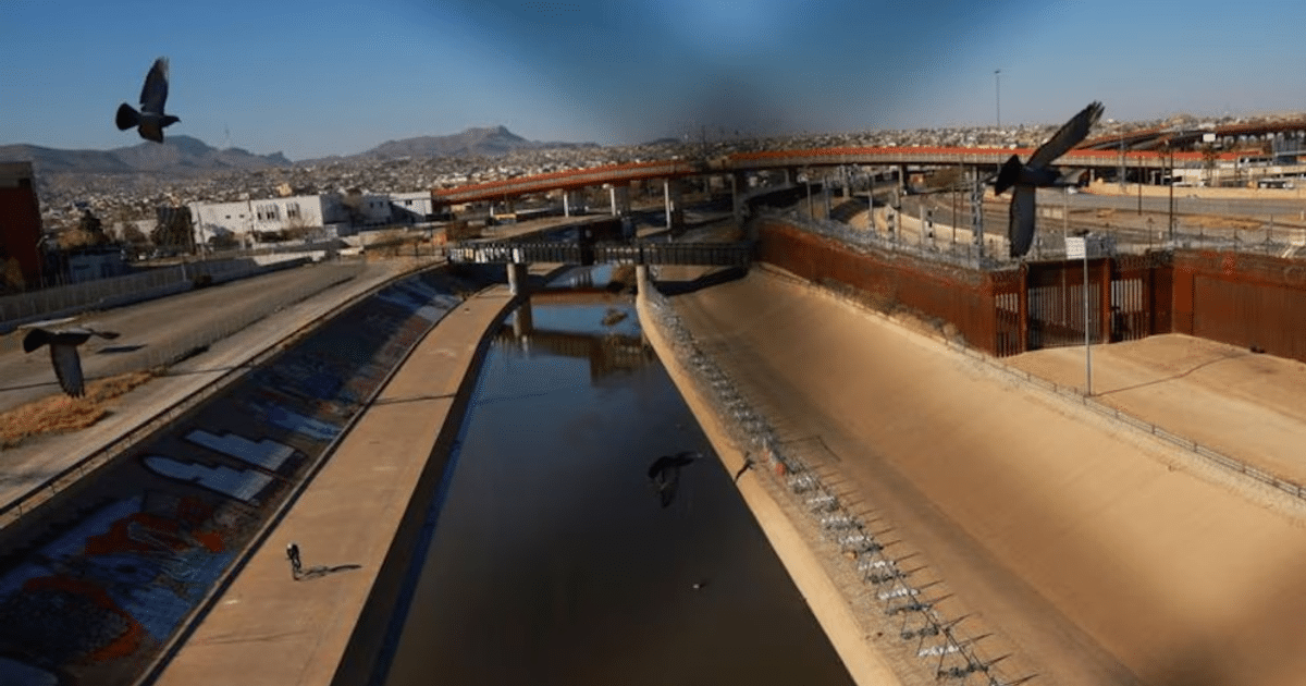 Paisaje urbano en la frontera de Texas con canal seco, valla fronteriza y aves en el cielo.