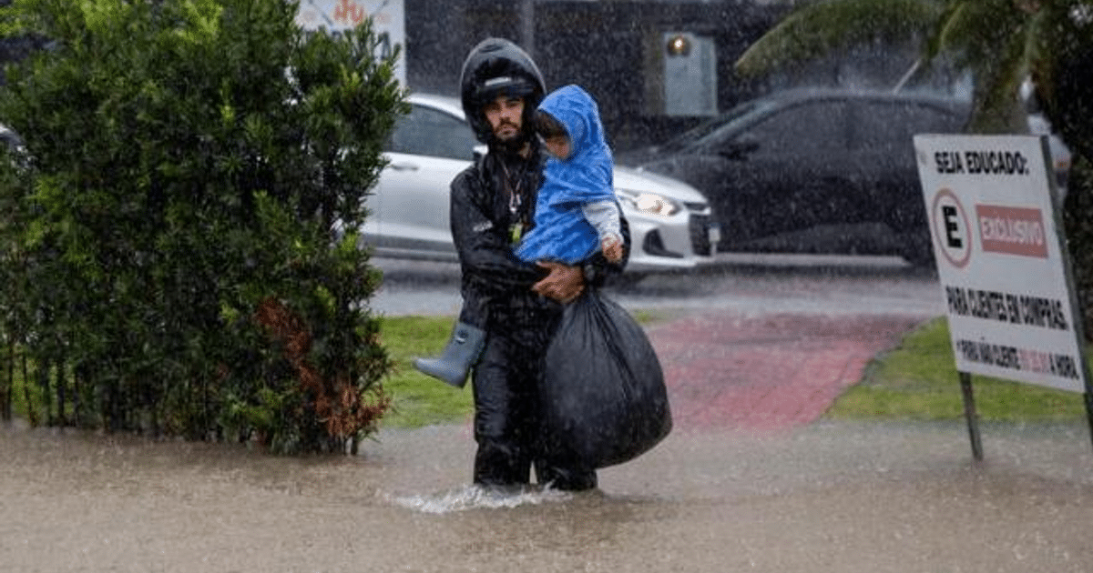 Hombre con casco y niño en brazos en área inundada de Santa Catarina