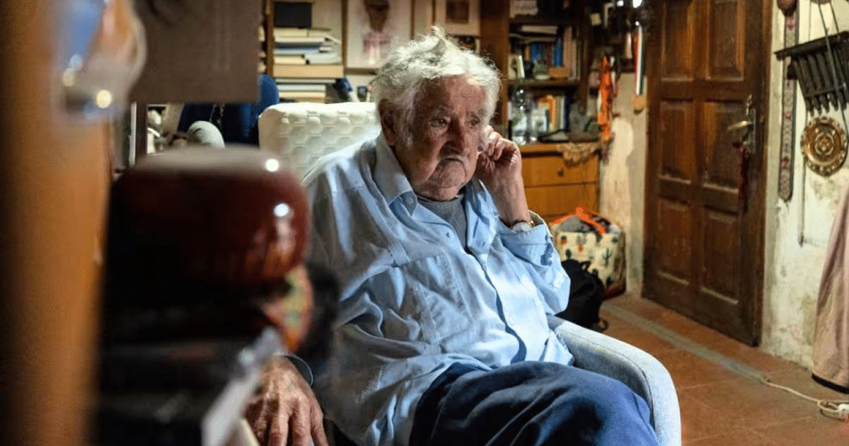 José Mujica sentado en un sillón, pensativo, en un entorno acogedor con estanterías y decoración.