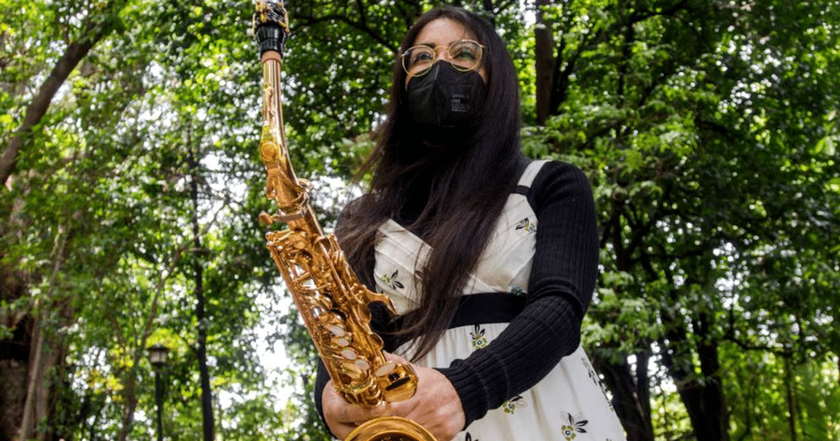 María Elena Ríos Ortiz con saxofón en un parque, vestida con atuendo floral y mascarilla negra.