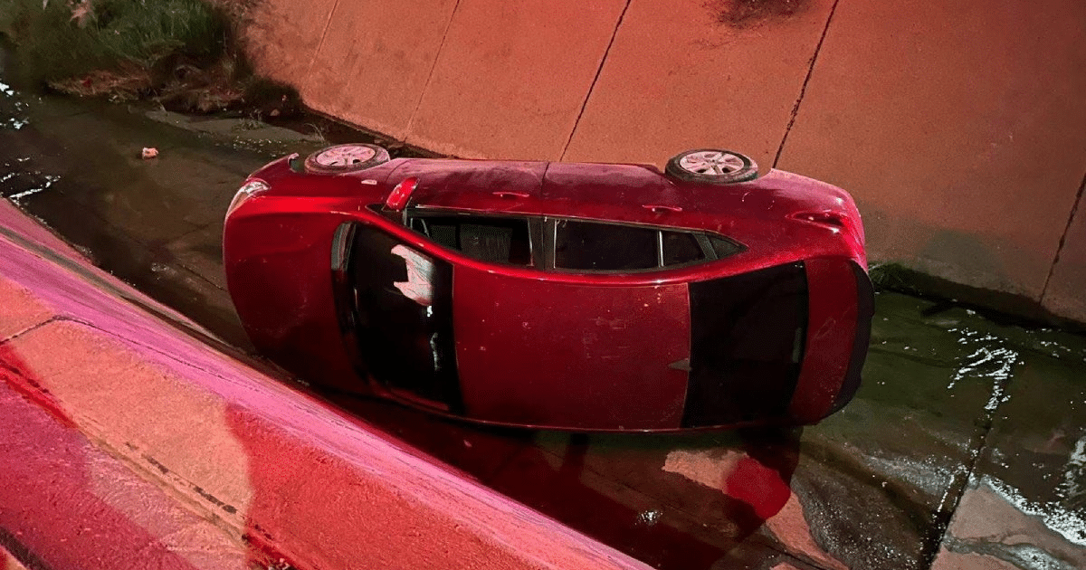 Automóvil rojo volcado en canal pluvial en Mazatlán, iluminado por luces de emergencia.