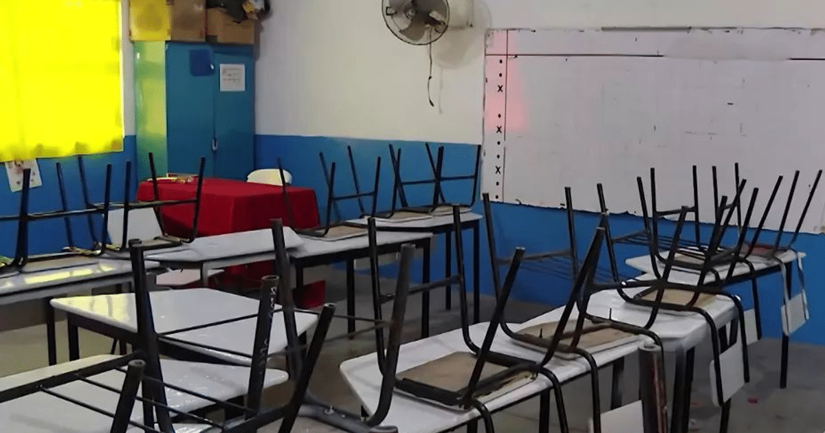 Aula vacía con mesas y sillas invertidas, ventilador en la pared, ventana con cortinas amarillas, y un pizarrón al fondo.