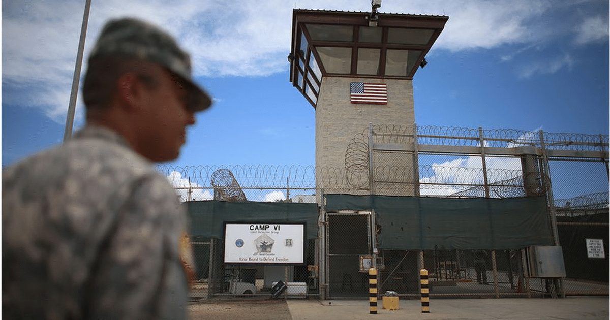 Torre de vigilancia y cercas de alambre de púas en Guantánamo, con un militar en primer plano y bandera estadounidense ondeando.