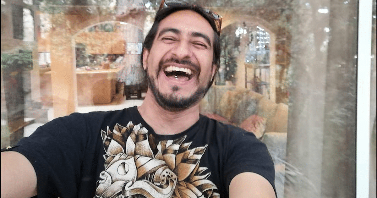 Persona sonriendo y tomando un selfie en un ambiente interior acogedor, con camiseta negra de diseño gráfico.