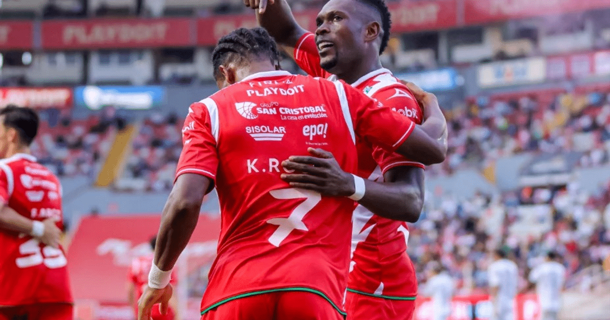 Dos jugadores de fútbol con uniformes rojos celebran en el campo, uno con el número 7, en un estadio lleno de espectadores.