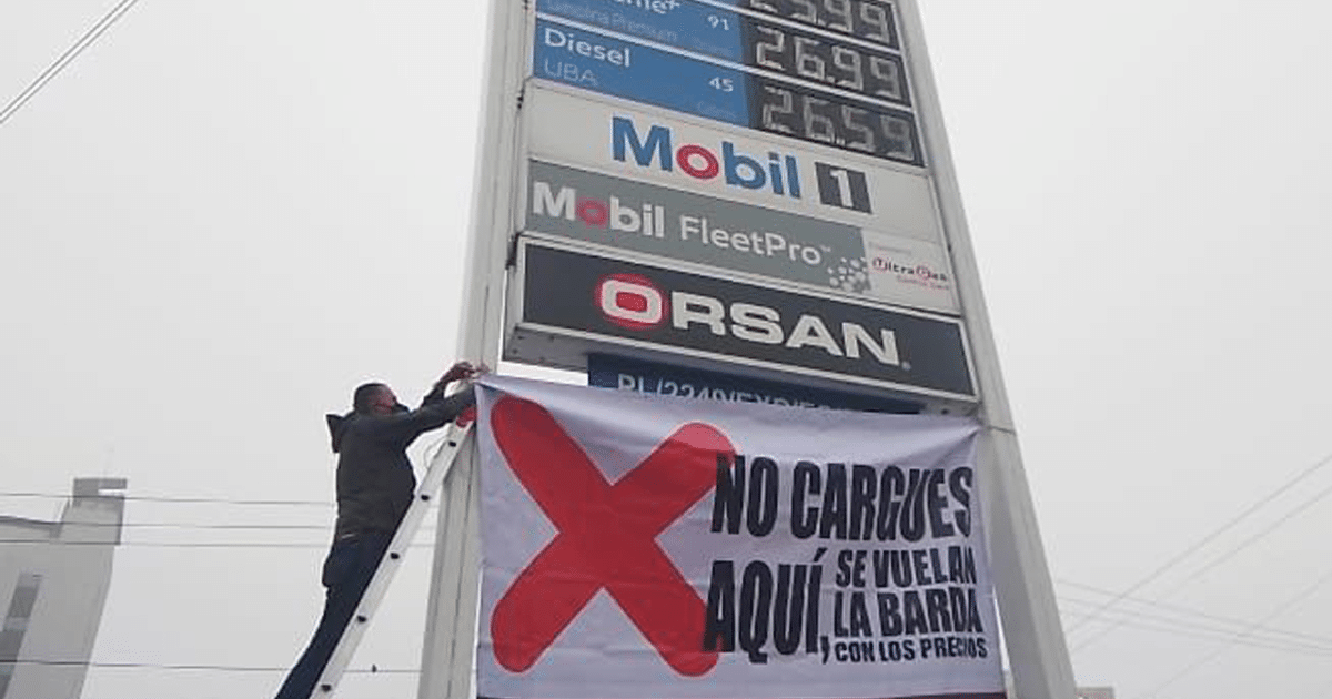 Persona colocando pancarta de advertencia en gasolinera con tablero de precios de combustible al fondo.