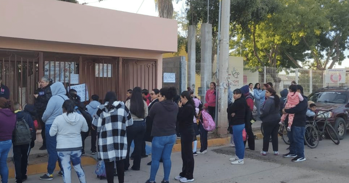 Grupo de personas, incluyendo niños, esperando frente a una escuela con mochilas y un vehículo estacionado, en un ambiente de manifestación.