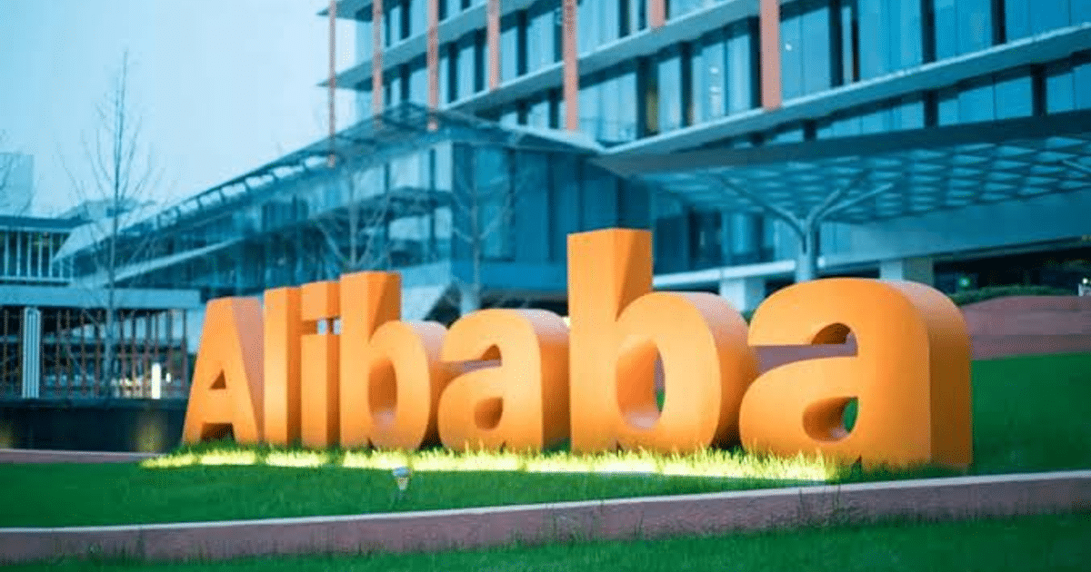Letrero de Alibaba en letras naranjas sobre césped con un edificio moderno al fondo.