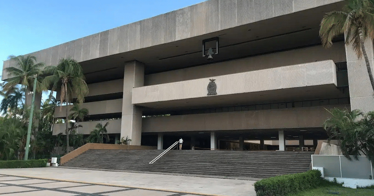 Edificio moderno con fachada de concreto, palmeras, campana y emblema en entorno tropical.