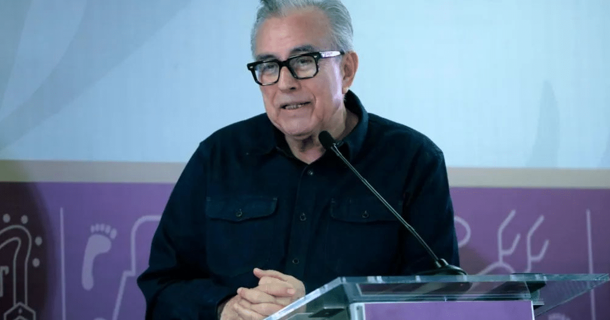 Gobernador Rubén Rocha Moya hablando en una conferencia de prensa con un micrófono en mano y un fondo decorativo.