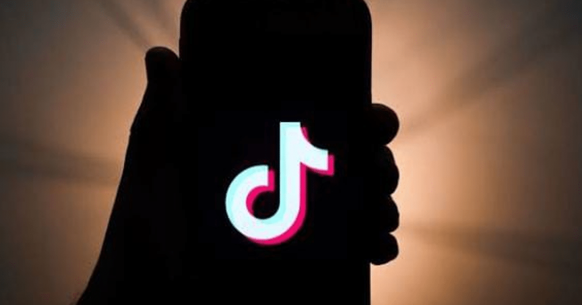 Mano sosteniendo un teléfono móvil con el logotipo de TikTok en la pantalla, sobre un fondo con sombras suaves.