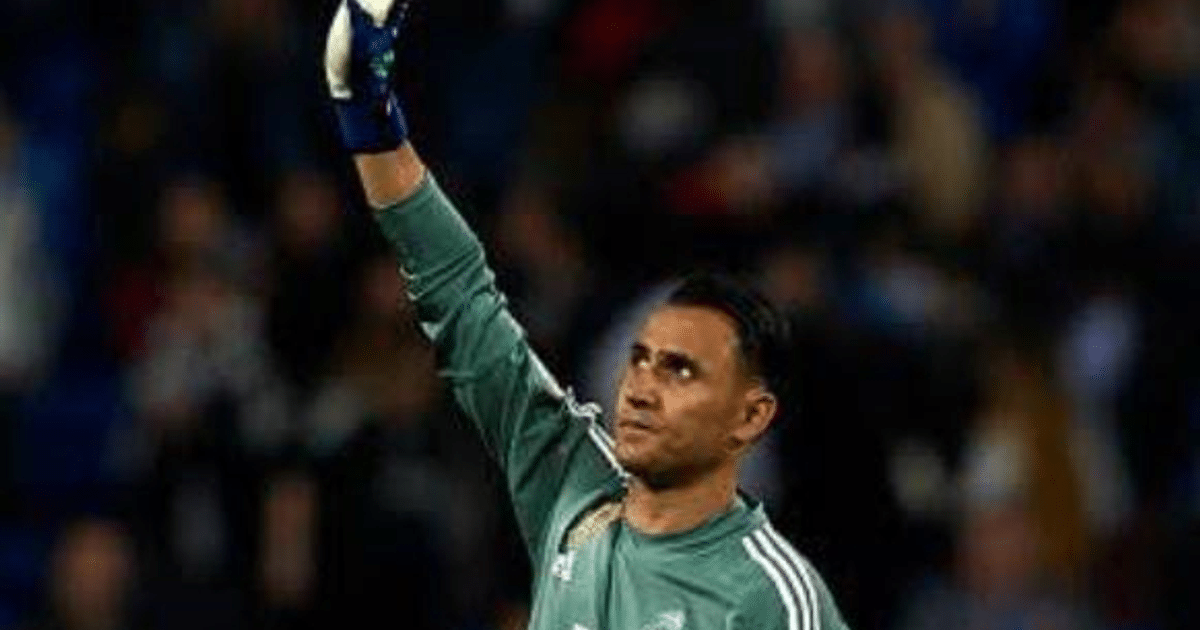 Portero de fútbol con camiseta verde levantando una mano en un estadio.