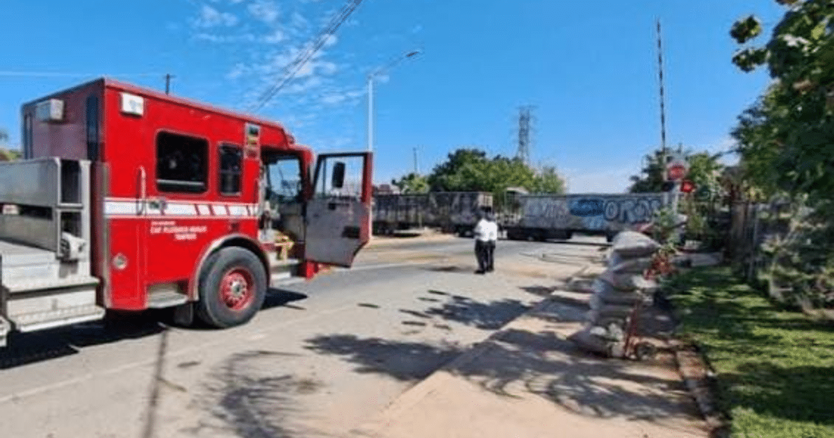 Camión de bomberos en accidente ferroviario en Culiacán con hombre dirigiendo tráfico y tren al fondo.