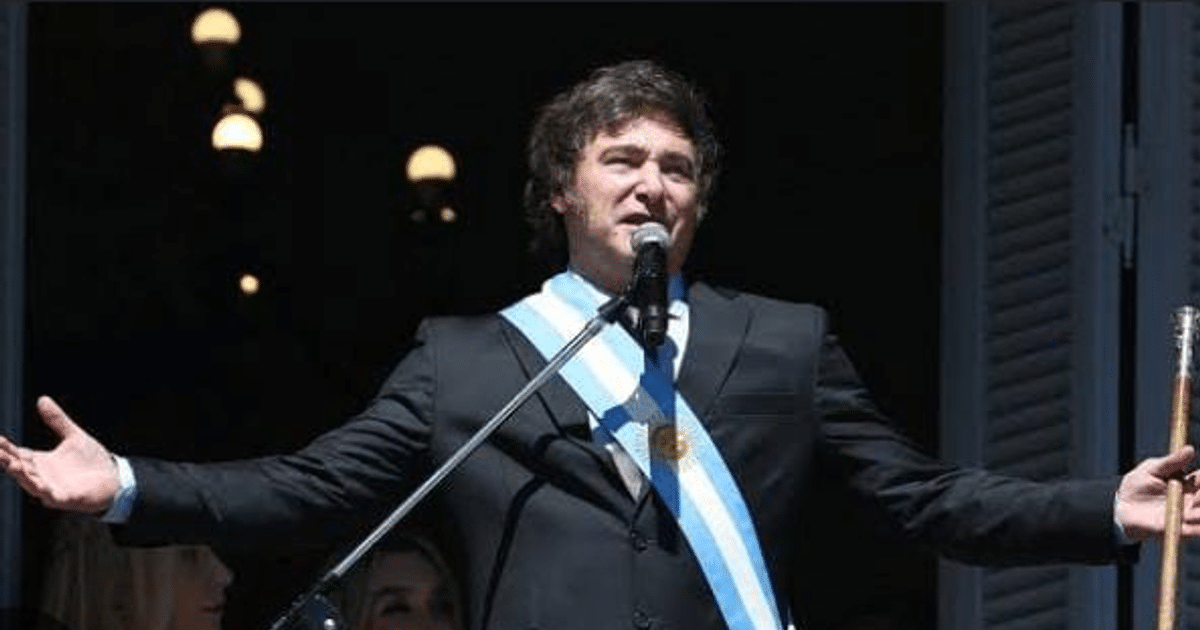 Javier Milei en traje negro con banda argentina hablando en un micrófono durante un evento público.