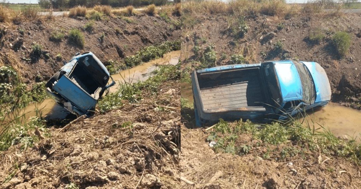 Camioneta azul volcada en canal de agua en Guasave, rodeada de vegetación y barro, con carretera al fondo.