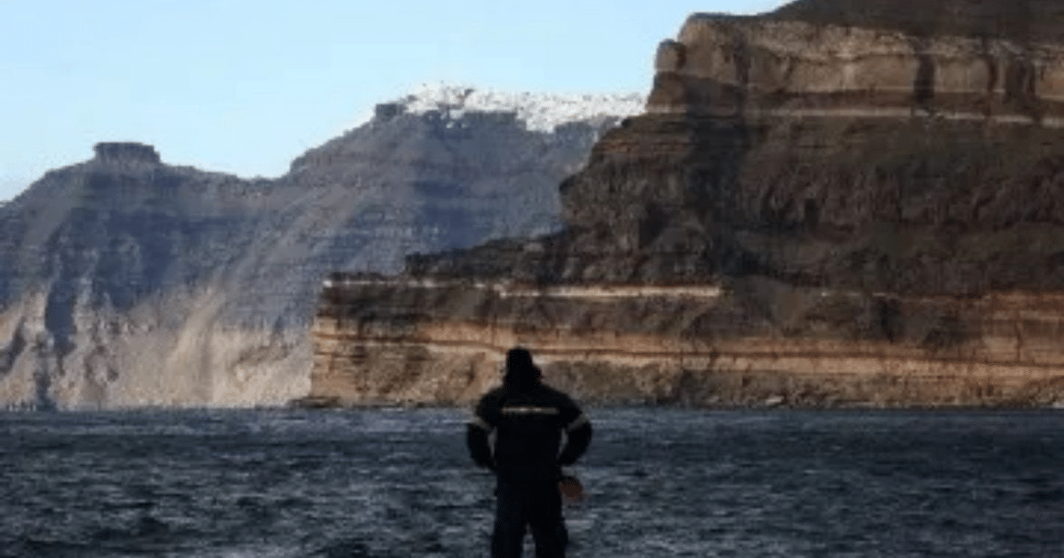 Hombre de espaldas frente al mar en un paisaje costero con acantilados y montañas en islas griegas durante actividad sísmica.