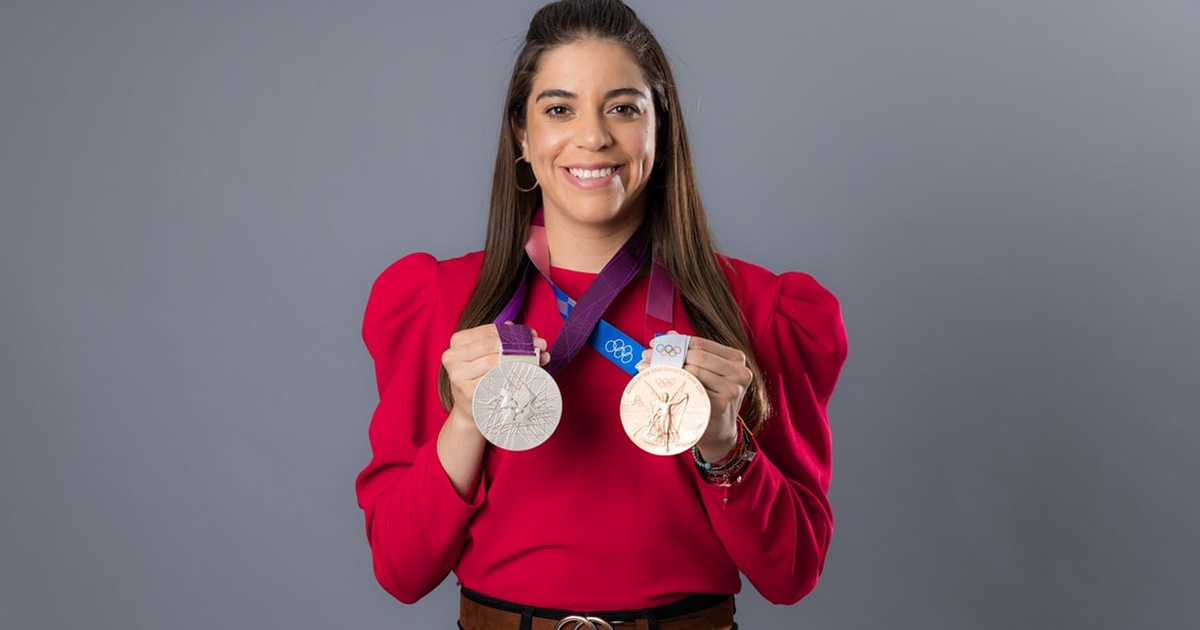Alejandra Orozco sosteniendo medallas olímpicas de plata y bronce, con una blusa roja y fondo gris.