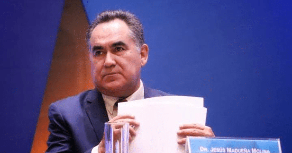 Hombre serio en traje oscuro con papeles en mano, fondo azul de conferencia