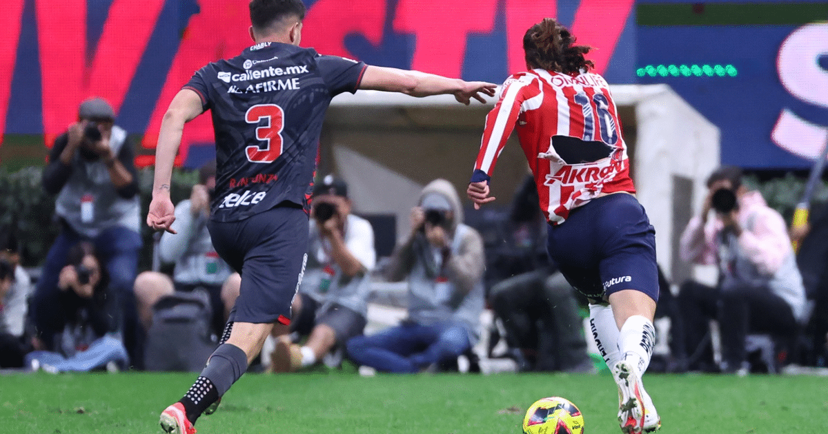 Dos jugadores de fútbol en acción durante un partido en el Estadio Akron, uno con uniforme oscuro número 3 y otro con uniforme a rayas rojas y blancas número 11.