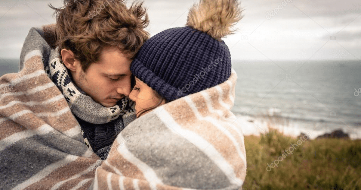 Pareja joven abrazada bajo una manta con el mar y cielo nublado de fondo