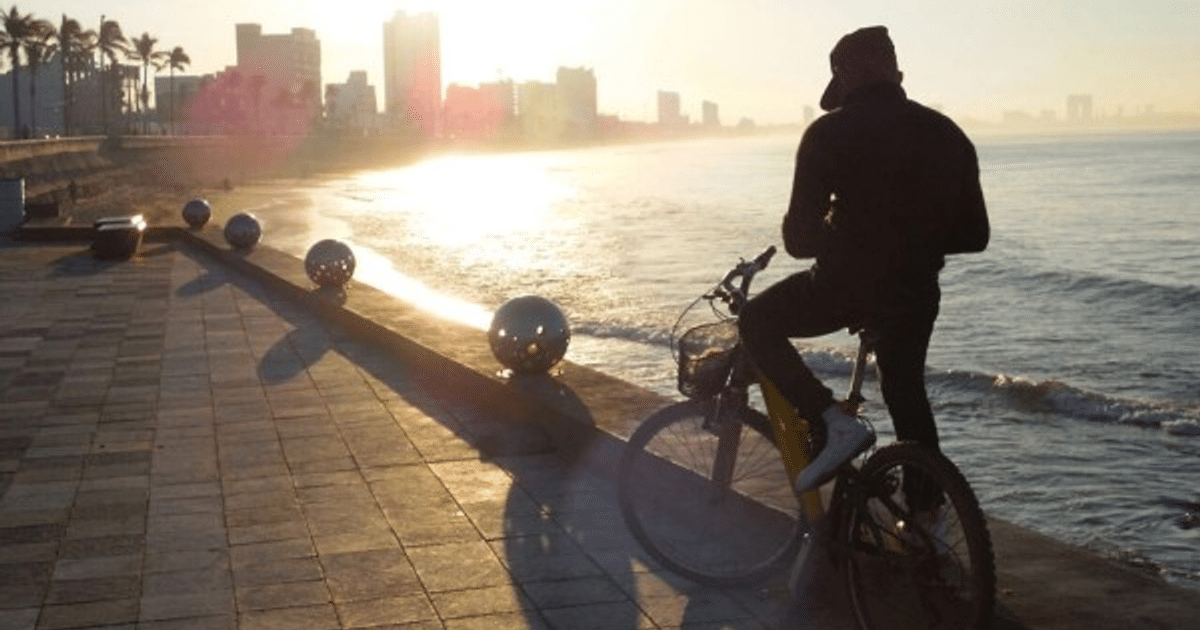 Persona en bicicleta en el malecón de Sinaloa al amanecer, con el mar y edificios de fondo.