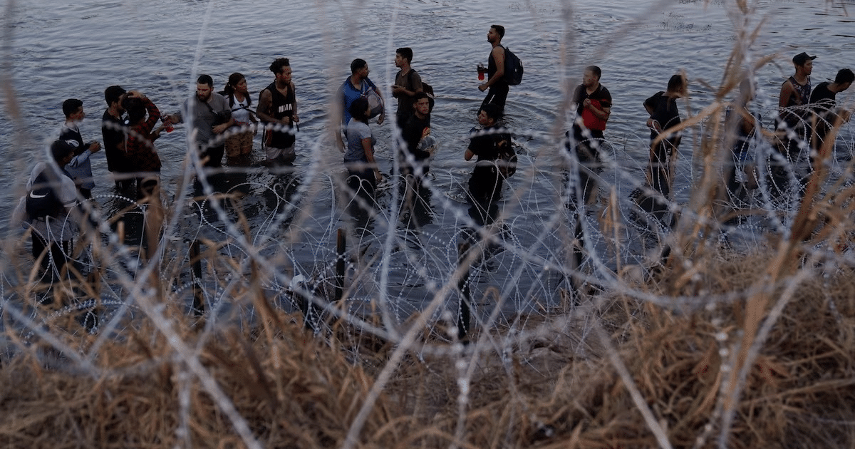 Grupo de personas en el agua cerca de alambre de espino, simbolizando un cruce fronterizo.