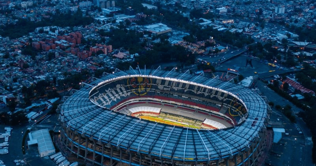 Vista aérea nocturna del Estadio Azteca iluminado, rodeado de edificios y calles urbanas.