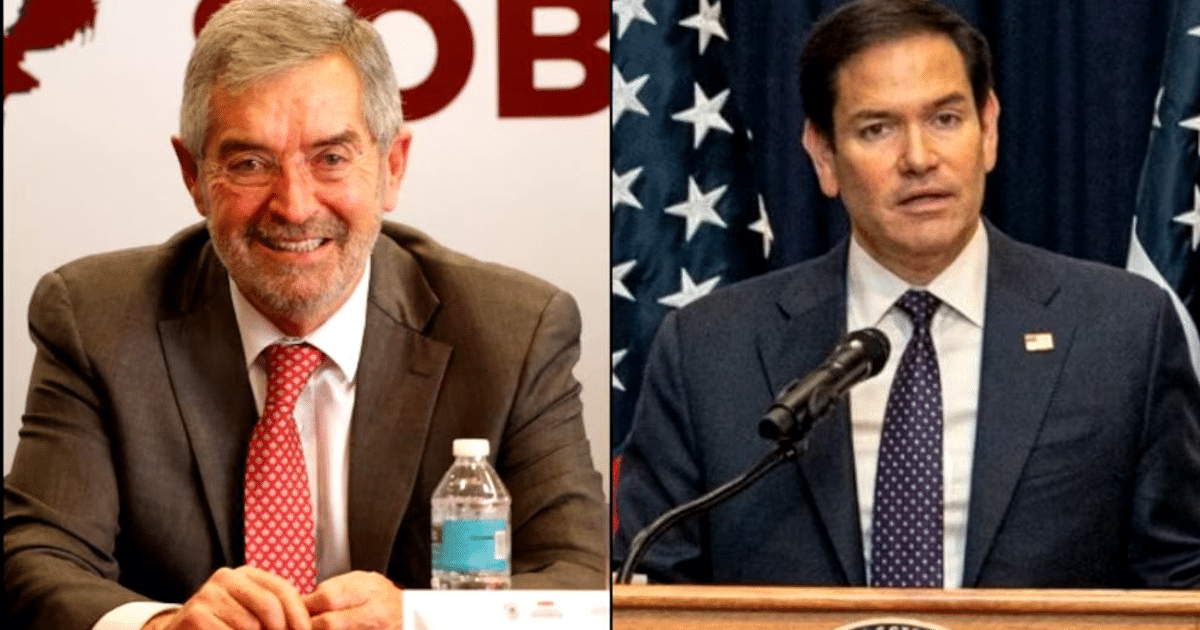 Dos hombres en un evento formal, uno sonriendo en traje gris y otro hablando en un podio con banderas de EE. UU. al fondo.