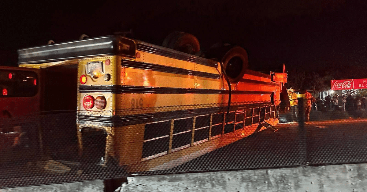 Autobús escolar volcado en carretera nocturna con luces reflejadas y personas al fondo.