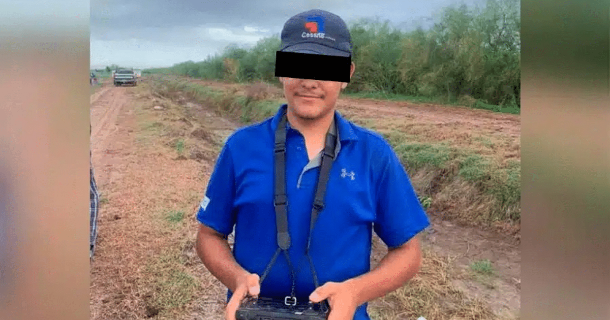 Persona con gorra y camiseta azul en un paisaje rural, sosteniendo un control remoto, con vehículos y vegetación al fondo.