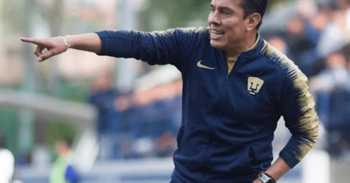 Entrenador de fútbol señalando en un estadio, vestido con chaqueta azul oscuro y detalles dorados.