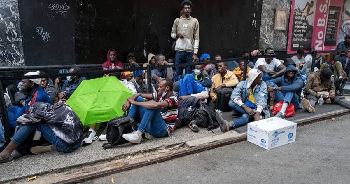 Grupo de migrantes sentados en la acera con mochilas y un paraguas verde, cerca del Hotel Roosevelt en Nueva York.